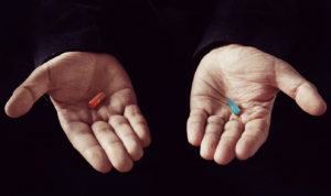 Two hands one holding a blue pill, the other holding a red pill
