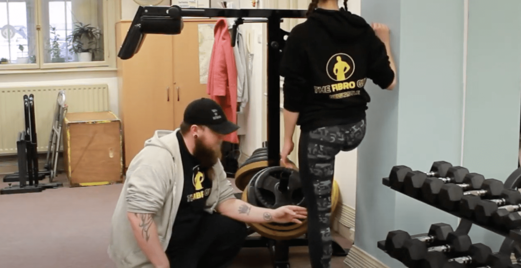A man helping a woman wearing gym kit do an ankles exercises up against a wall.