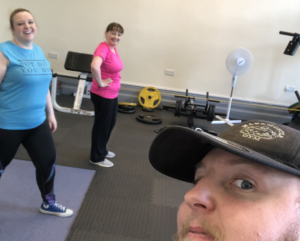 A man with a black hat smiles, with two women in the background, one with a pink top, the other with a blue top.