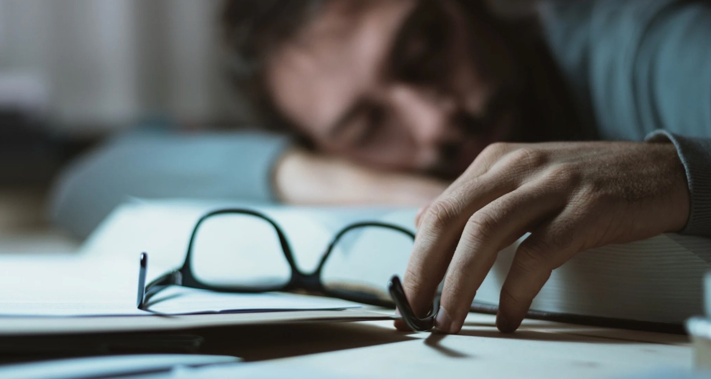 Tired man slumped on a desk with glasses removed, unable to sleep with POTS