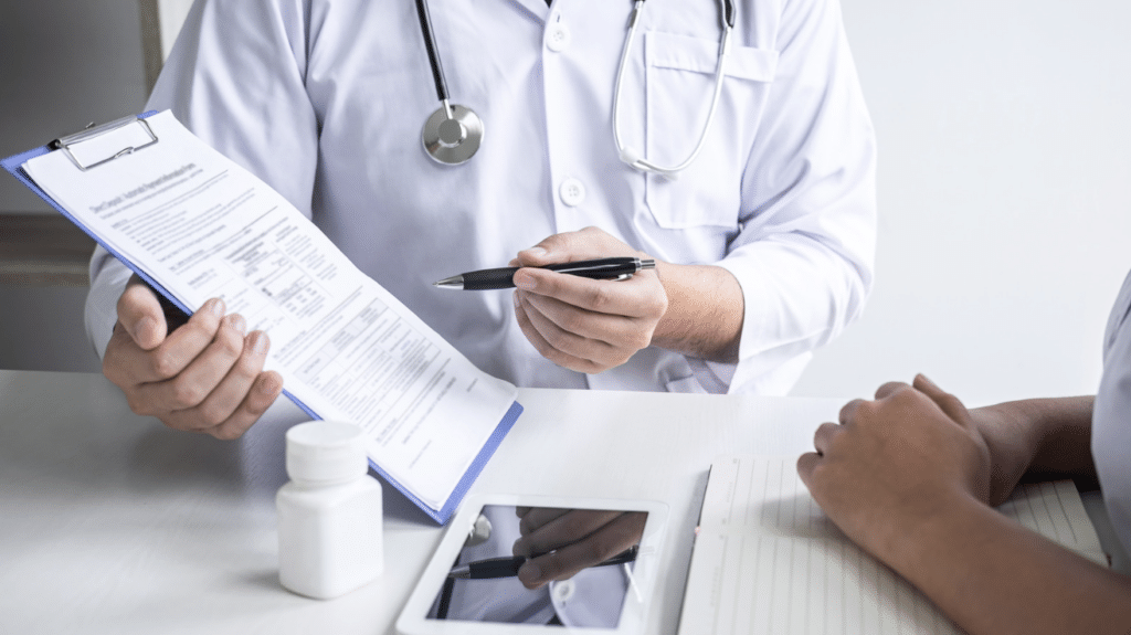 Clinician in a white coat holding a clipboard and pen across a desk from someone at an appointment