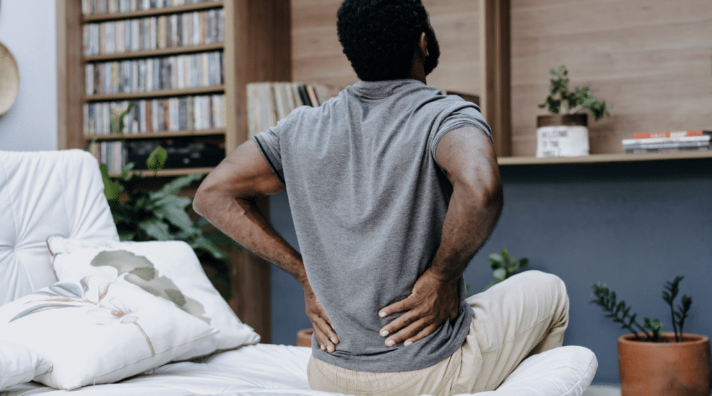 A man sitting on the edge of a bed with both hands pressed into his lower back