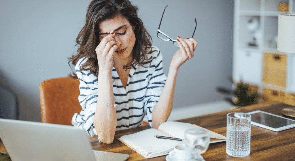 A woman at a desk with a laptop, eyes closed, pinching the bridge of her nose